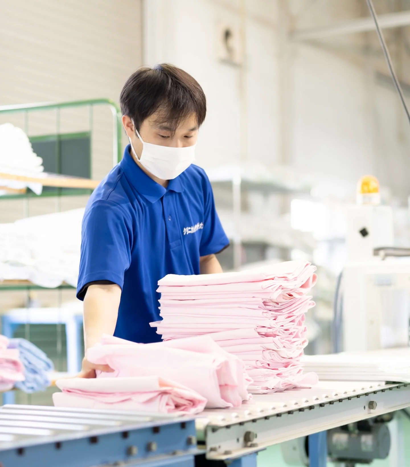 作業員が工場のコンベヤー横で淡いピンク色のリネンを整えながら積み重ねている。